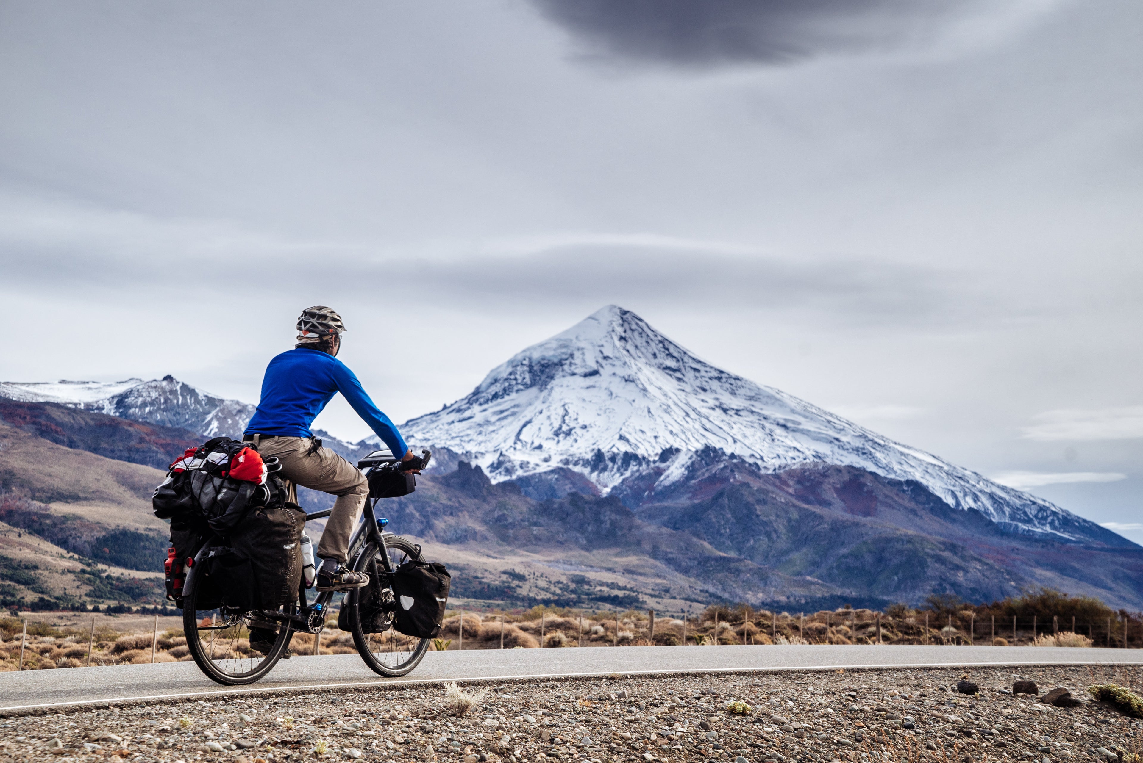 A rider on a Pinion-equipped touring bike, loaded with panniers riding on a road near a tall mountain.